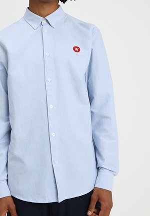 Light blue long-sleeve button-up shirt with a red oval logo patch featuring white initials on the left chest.