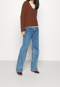 Hnědý pletený svetr s výstřihem do V navlečený na světle šedém topu, zkombinovaný s modrými širokými džínami a hnědými podpatkovými mulemi.