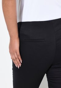 Schwarze Hose mit glattem Textur, ausgestattet mit einer diskreten Gesäßtasche und einer taillierten Passform. Eine Hand liegt auf dem Stoff.