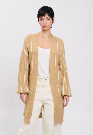 Person trägt einen beige-weiß gestreiften, übergroßen Cardigan, weißes Top, cremefarbene Hose und eine einfache goldene Halskette vor einem hellgrauen Hintergrund.