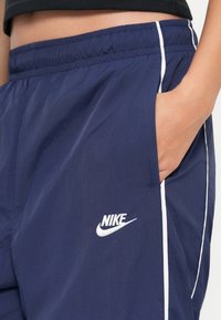 Artimiausias kadras rodo žmogaus ranką kišenėje, vilkinčioje tamsiai mėlynas Nike sportines kelnes su baltomis juostelėmis ir logotipu ant šlaunies.