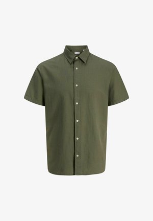 Adolescent aux cheveux bouclés souriant, portant une chemise boutonnée à manches courtes vert olive, un pantalon blanc et des baskets beige.