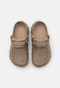 Beige clogs made of soft, molded plastic with ventilation holes and a fabric strap. Features a textured footbed for comfort.