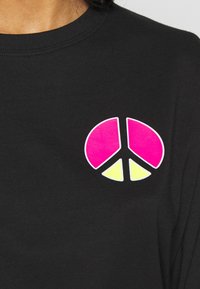 Schwarzes Langarmshirt mit einem bunten Friedenssymbol-Graphic. Das Symbol verwendet die Farben Pink und Gelb und ist auf einem glatten Stoff aufgebracht.