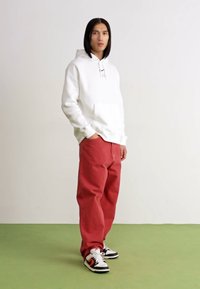 Felpa bianca con cappuccio, pantaloni rossi larghi e sneakers nere con dettagli arancioni. Design semplice con tasca frontale sulla felpa.