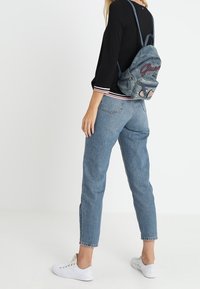 Mochila de mezclilla azul con detalles bordados, correas para los hombros y superficie texturizada. Combinada con jeans azul claro y zapatillas blancas.