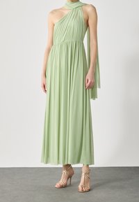 Femme portant une robe maxi fluide vert clair sans manches avec un col halter asymétrique et un détail d'écharpe nouée. Elle porte des sandales à talons hauts dorées.
