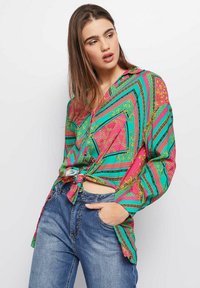 Camisa de manga larga colorida con un patrón geométrico en rosa, verde y negro, con acentos dorados, atada en la cintura. Combinada con jeans de mezclilla.