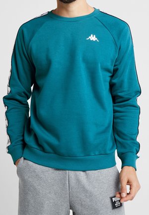 Man som bär en teal sweatshirt med vita logotyper över bröstet och på ärmarna, ihop med ljusgråa sweatpants med svart lappar.