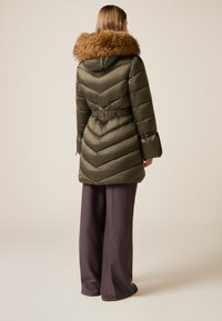 Abrigo acolchado verde oliva con capucha con ribete de piel, costuras en chevron, cintura ceñida y mangas anchas, combinado con pantalones grises de pierna ancha.