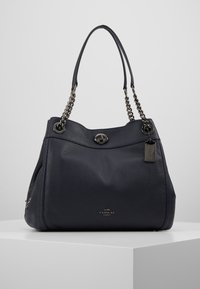 Sac à main Coach en cuir noir avec anses en chaîne argentée, fermeture à tourniquet et étiquette logo en métal sur un piédestal blanc.