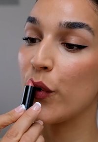 Läppstift i ett svart rör som appliceras på läpparna. Läpparna har en dämpad rosa nyans, med en matt finish och en lätt textur.