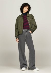 Chaqueta bomber acolchada en verde oliva con puños de canalé, suéter de cuello alto con rayas en azul marino y rojo, pantalones de pierna ancha grises de talle alto y zapatos blancos.