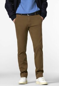 Man wearing brown pants, white sneakers, blue shirt, navy jacket, hands in pockets, standing against a plain light background.