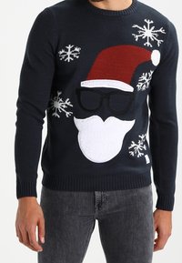Pull bleu marine avec un grand visage du Père Noël portant des lunettes et un chapeau rouge, entouré de flocons de neige blancs, porté par une personne en jean gris foncé.