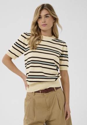 Nő bézs, barna és világoskék csíkos rövid ujjú pulóverben, pliszírozott bézs nadrággal és barna övvel, egyik kezét csípőjén tartva pózol.