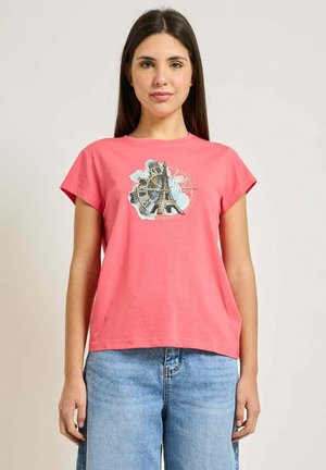 Donna che indossa una maglietta corallo con grafica della Torre Eiffel e orologio, abbinata a jeans azzurri, in piedi davanti a uno sfondo semplice.