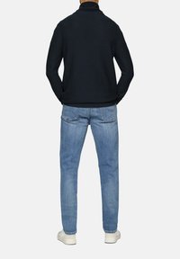 Marinefarbener Pullover mit hohem Kragen, kombiniert mit hellblauen Jeans. Weiße Sneakers sind unten sichtbar. Schlichtes, figurschmeichelndes Design.