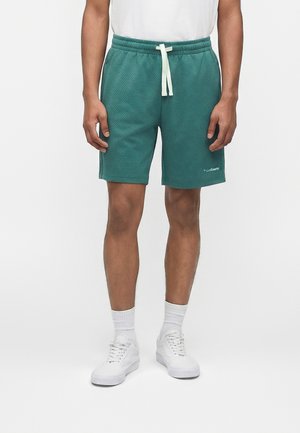 Man wearing teal textured shorts with white drawstring, white socks, and white sneakers, standing against a plain background.