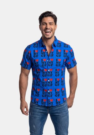 Glunderende man met donker haar draagt een felblauw korte mouwen shirt bedekt met rode harten en zwarte tekst "I love my GF", gecombineerd met blauwe spijkerbroek.