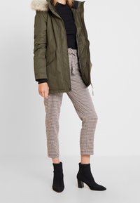 Parka verde oliva con cappuccio in pelliccia sintetica, abbinata a pantaloni cropped beige a quadri e stivali neri alla caviglia. Tessuto liscio e vestibilità su misura.