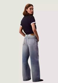 Jeans sbiaditi blu a gamba larga con vita alta, dotati di due tasche posteriori e una toppa in pelle marrone. Abbinati a una maglietta nera aderente.