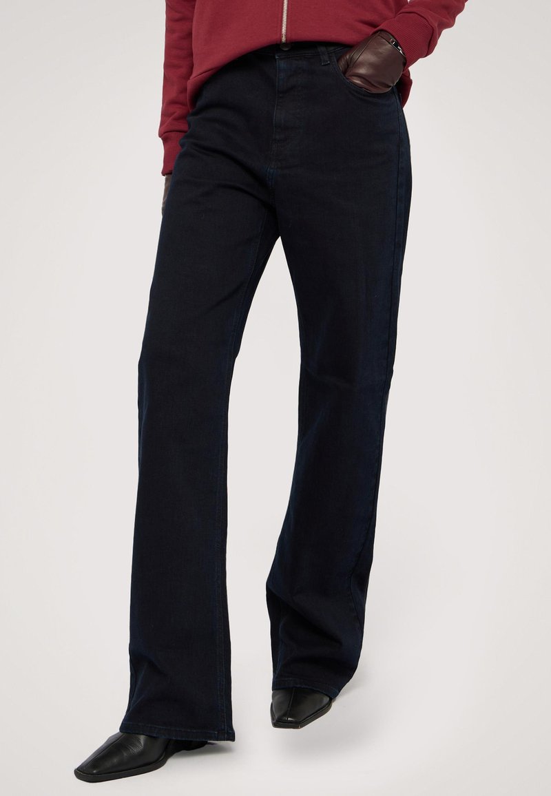 Tmavě modré džíny rovného střihu vyrobené z denimu, s kapsami a vysokým pasem. Vybavené černými botami s špičatou špičkou.