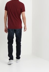 Bordový polo tričko s krátkým rukávem v kombinaci s tmavě modrými džíny a světle šedými teniskami, pohled zezadu na jednobarevném světlém pozadí.