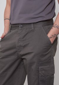 Pantaloni cargo grigi con chiusura a bottone, tasche frontali e una tasca laterale. Presentano una texture liscia e un design aderente.