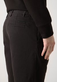 Pantalons en coton noirs avec une coupe slim, poches latérales et deux poches arrière avec un détail d'étiquette. Le tissu semble lisse et texturé.