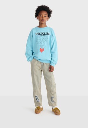 Kind draagt een lichtblauwe trui met de tekst "PICKLES" en een groenteafbeelding, beige broek met blauwe stukken en bruine schoenen met gele veters.