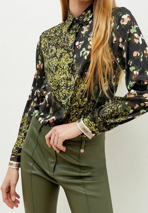 Kobieta w długim rękawie, koszula na guziki z abstrakcyjnym kwiatowym wzorem w zielono-czarno-beżowych kolorach, w połączeniu z oliwkowozielonymi spodniami z wysokim stanem.