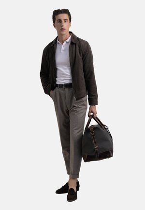 Mann in braunem Jackett, weißem Poloshirt, grauen Hosen und Slippern, der eine dunkelbraune Leder- und Stoffreisetasche hält und vor einem weißen Hintergrund steht.