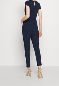 Granatowy kombinezon z krótkimi rękawami, detalami w kształcie dziurki z tyłu, dopasowanymi spodniami oraz świecącymi srebrnymi sandałami na wysokim obcasie. Gładka faktura materiału.
