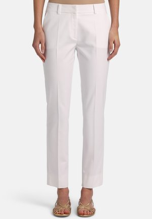 Ženska, oblečena v bele hlače po meri z zlikanimi pregibi, stranskimi žepi in bež sandale z odprtimi prsti ter peto, stoji ob preprostem ozadju.