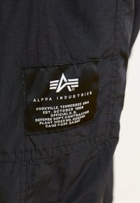 Étiquette en tissu noire avec texte et logo blancs, indiquant "ALPHA INDUSTRIES" et détaillant la date de création de l'entreprise ainsi que des identifiants supplémentaires.