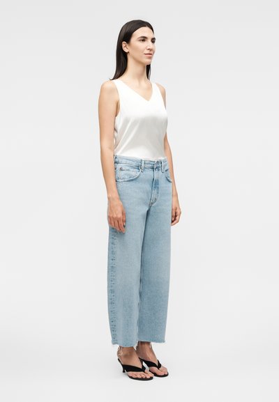 Γυναίκα με μακριά σκούρα μαλλιά, φορά λευκό αμάνικο τοπ με V λαιμόκοψη, ανοιχτούς μπλε τζιν παντελόνια με φαρδιά μπατζάκια και μαύρα πέδιλα με τακούνι, όρθια πλευρικά.