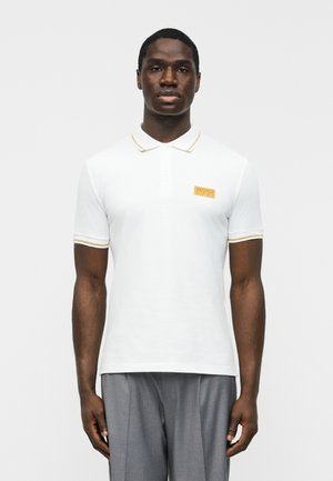 Mustanahasiline mees, kes kannab valget lühikeste varrukatega polosärki kuldse äärega ja halli pükse, seisab ühevärvilise heledana tausta ees.