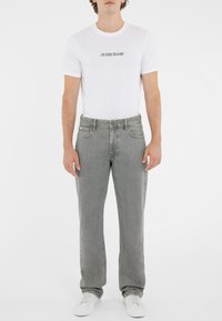 Balta marškinėliai su "GUESS JEANS" logotipu, derinami su šviesiai pilkomis tiesiomis kelnėmis. Modelis dėvi baltus sportbačius. Lygus audinys, laisvalaikio stilius.