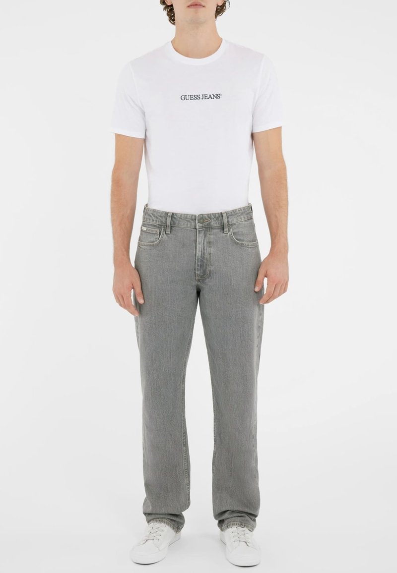 Balta marškinėliai su "GUESS JEANS" logotipu, derinami su šviesiai pilkomis tiesiomis kelnėmis. Modelis dėvi baltus sportbačius. Lygus audinys, laisvalaikio stilius.