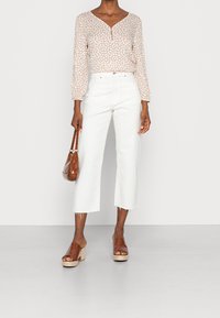 Blusa beige a maniche lunghe con motivo astratto bianco, scollatura a V e bottone. Jeans bianchi a vita alta e gamba ampia. Sandali con tacco marroni e borsa.