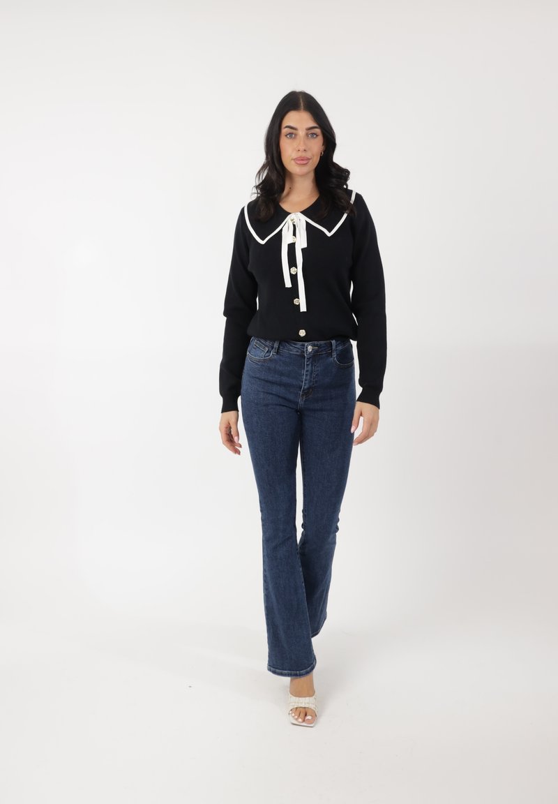 Suéter negro con gran cuello blanco y botones en el frente, combinado con jeans de campana azules. La modelo lleva sandalias de tiras en color claro.