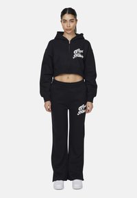 Donna che indossa un cropped hoodie nera con zip e pantaloni della tuta abbinati con la scritta bianca "Good For Nothing", abbinati a sneakers bianche, in piedi di fronte.