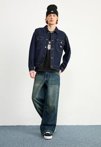 Mørk denimjakke med to forlommer, blå slidte baggy jeans og sort beanie. Sættet har en grafisk sort t-shirt under.