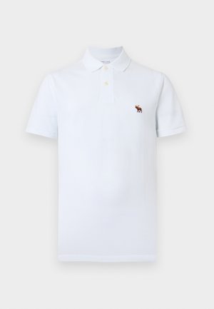 Fehér, rövid ujjú galléros póló két gombbal, melynek bal mellrészén egy kis barna jávorszarvas hímzett logó található.