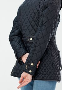 Black quilted jacket featuring a fitted design, gold hardware accents, and a belted waist. Visible stitching creates a diamond pattern.