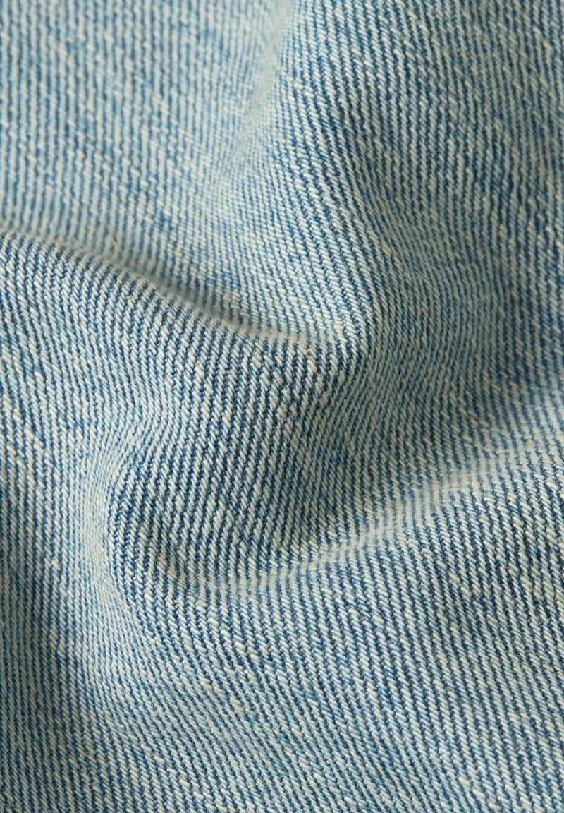 Primo piano di tessuto in denim azzurro con trama intrecciata visibile e pieghe sottili che creano ombre morbide.