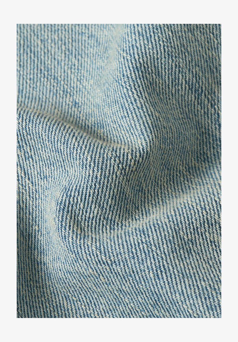 Primo piano di tessuto in denim azzurro con trama intrecciata visibile e pieghe sottili che creano ombre morbide.