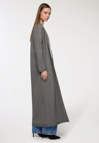 Długi, szary kardigan wykonany z miękkiego materiału, z szerokimi rękawami i luźnym krojem, w zestawieniu z jasnoniebieskimi dżinsami i czarnymi butami na obcasie.