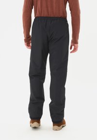 Pantaloni impermeabili neri con vita elastica, gambe affusolate e orli rinforzati, visti da dietro. Abbinati a scarpe marroni.